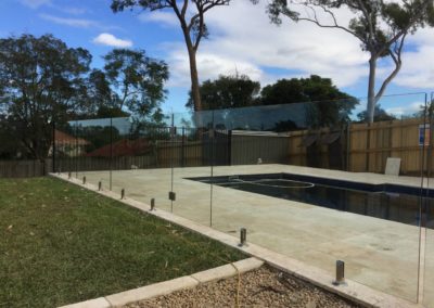 Backyard pool secured with frameless glass fencing, featuring stainless steel spigots on a landscaped lawn.