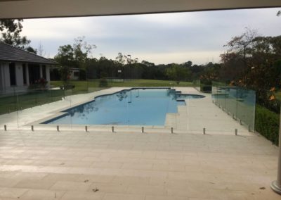 Spacious outdoor pool area enclosed with frameless glass fencing, viewed from a paved patio, creating an open and inviting space.