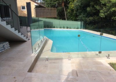 Frameless glass pool fence with square stainless steel spigots on travertine paving, surrounding a large pool beside a forested area.