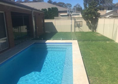 Frameless glass pool fence with stainless steel spigots along a clear blue pool, adjacent to a home and backyard lawn