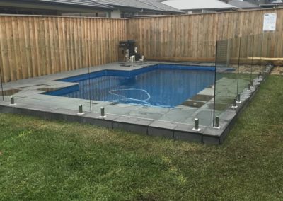 Elegant frameless glass pool fencing with stainless steel spigots, surrounding a blue tiled pool in a backyard with timber fencing.