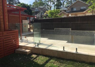 Frameless glass pool fence with polished stainless steel spigots on raised travertine pool area in a residential backyard.