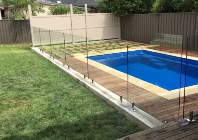 Standard and custom frameless glass pool fence panel along stairs for compliance, bordering a blue fibreglass pool with wooden deck.