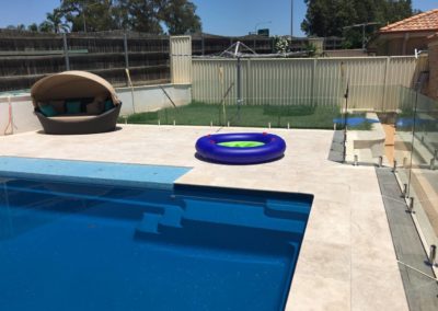 Crystal clear pool with frameless glass fence and a vibrant blue inflatable ring on the travertine surrounds.
