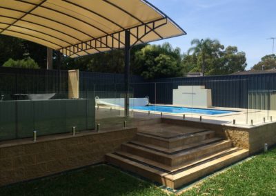 Frameless glass pool fence and glass balustrade core drilled through a sandstone block wall, with an outdoor awning covering a lounge area